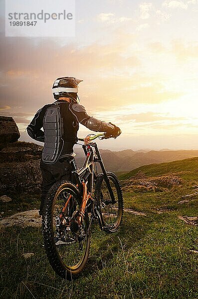 Ryder in voller Schutzausrüstung auf dem mtb von zwei Anhänger Fahrräder reitet entlang der grünen Gras in den Strahlen des Sonnenuntergangs Sonne vor dem Hintergrund einer Landschaft von felsigen Gelände unter der untergehenden Sonne und Wolken