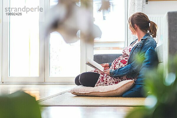 Schwangere kaukasische glückliche Mutter sitzt auf dem Boden und liest ein Buch