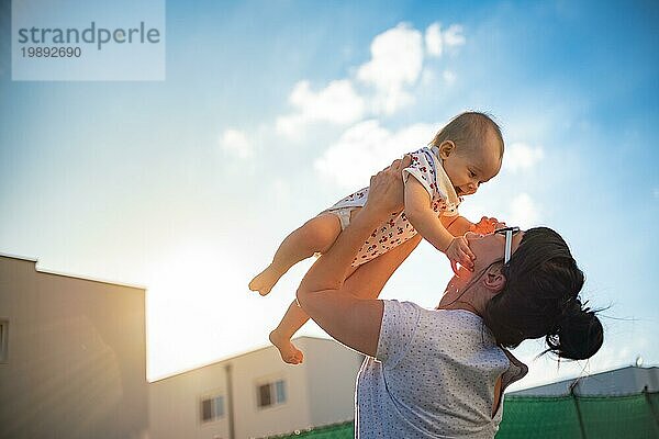 Ein emotionales Bild von 1 Jahr altes Baby und ihre Mutter hält sie in die Luft gegen blaün Himmel und helle Sonne. Mutterschaft Konzept