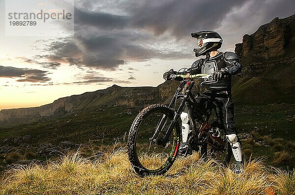 Der Fahrer in voller Schutz auf einem Mountainbike steht auf einem Hügel und schaut auf den Sonnenuntergang auf dem Hintergrund der Felsen im Kaukasus