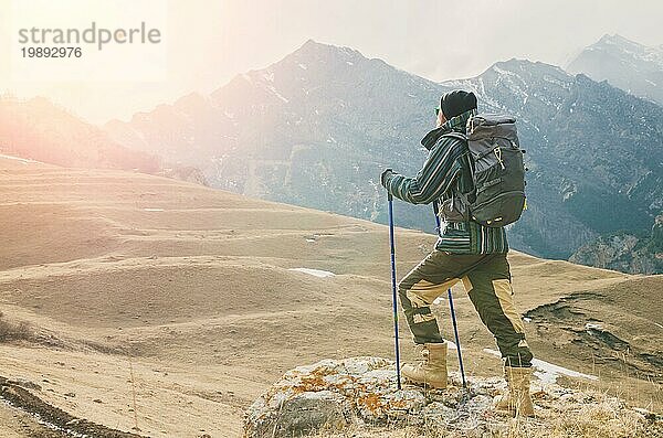 Ein Mann mit Bart und Sonnenbrille in einer Membranjacke  Hut  mit einem Rucksack und Stöcken für Nordic Walking  ein Reisender  der in der freien Natur steht und auf die Berge schaut