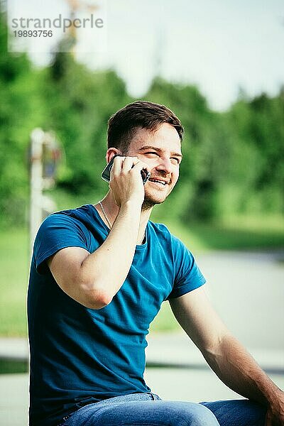 Hübscher junger Mann sitzt im Freien und spricht mit jemandem am Telefon