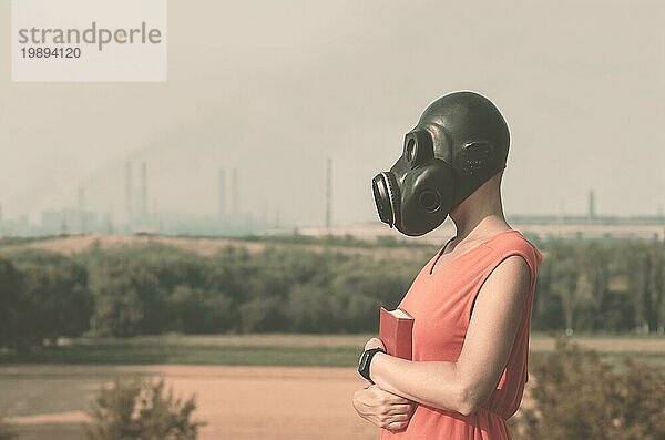 Junges Mädchen mit Gasmaske in einem roten Kleid und einem Buch in den Händen vor dem Hintergrund rauchender Fabrikschornsteine
