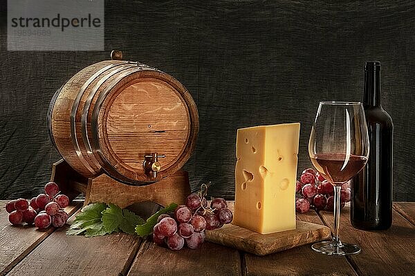 Ein Glas Rotwein mit einer Flasche  ein Weinfass  ein großes Stück Käse  Trauben und Weinblätter  Seitenansicht auf einem dunklen rustikalen Hintergrund  Low Key Foto mit einem Platz für Text