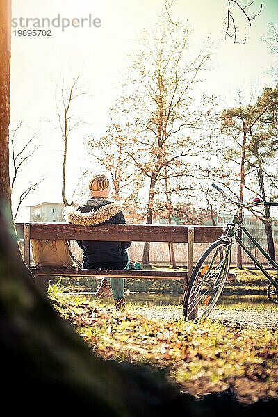 Rückenansicht einer jungen Frau mit Fahrrad  die die Sonne auf einer Parkbank genießt  Frühling