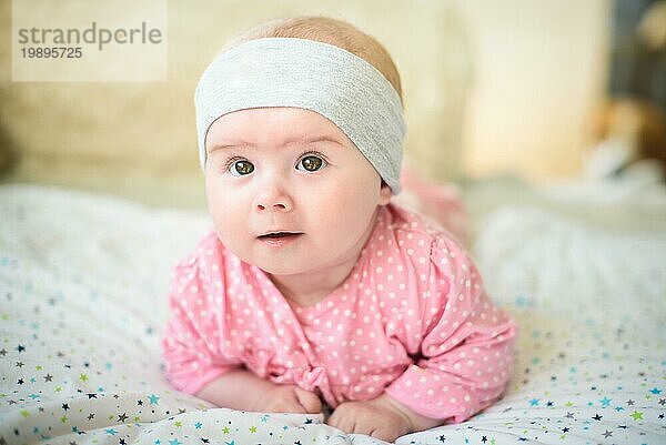 Cute 6 Monate alt Baby Mädchen Kleinkind auf einem Bett auf dem Bauch mit dem Kopf nach oben in die Kamera mit ihren großen Augen. Natürliches Schlafzimmer Licht
