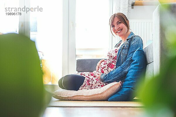 Schwangere kaukasische glückliche Mutter sitzt auf dem Boden  berührt ihren Bauch  blumiges Kleid