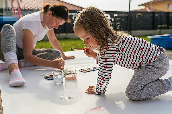 Focused Kind mit Mutter Malerei mit Plakatfarbe im Freien auf dem Boden auf großen Blatt Papier. Kreative Zeit verbringen mit Kindern Konzept