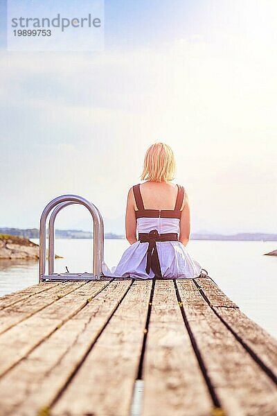 Junges Mädchen mit weißem Kleid sitzt auf einem Steg und genießt die Aussicht auf den See