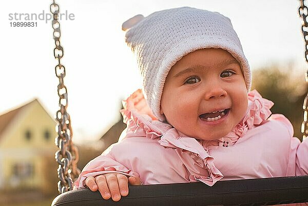 Adorable Baby Mädchen mit großen schönen Augen und eine Mütze mit Spaß auf einer Schaukel Fahrt auf einem Spielplatz in einem sonnigen Park