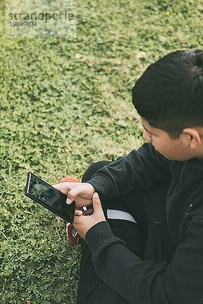 Draufsicht junger Millennial Junge sitzt im Park  in grünem Scutch Gras mit schwarzer Freizeitkleidung  schaut auf sein Smartphone  selektiver Fokus  Konzept der Technologie
