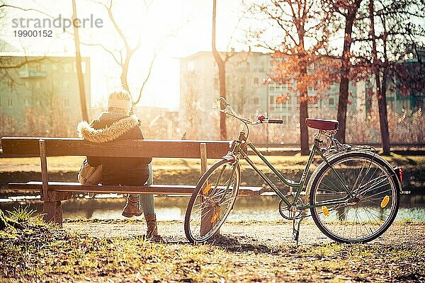 Rückenansicht einer jungen Frau mit Fahrrad  die die Sonne auf einer Parkbank genießt  Frühling