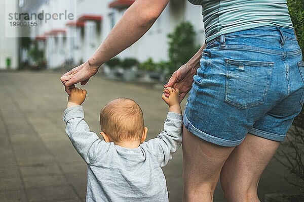 Konzept des Laufenlernens: Ein Kleinkind hält die Hand seiner Mutter und macht seine ersten Schritte. Nahaufnahme von hinten