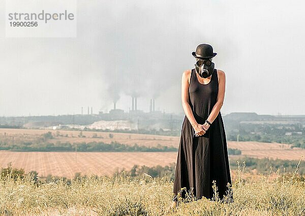 Junges Mädchen in einem schwarzen Kleid und einer Gasmaske vor dem Hintergrund rauchender Fabrikschornsteine