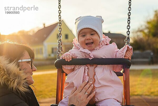 Adorable Baby Mädchen mit großen schönen Augen und eine Mütze mit Spaß auf einer Schaukel Fahrt auf einem Spielplatz in einem sonnigen Park