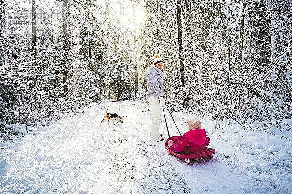 Mutter  die ihr Baby auf einem Schlitten durch den Winterwald zieht. Thema verschneiter Wald