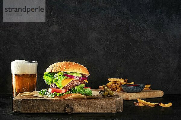 Burger und Bier. Hamburger mit Rindfleisch  Käse  Zwiebel  Tomate und grünem Salat  eine Seitenansicht auf einem dunklen Hintergrund mit Gurken und Pommes frites  mit einem Platz für Text. Selektiver Fokus