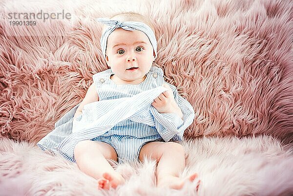 Nettes lächelndes Kind auf einem rosa Sofa. Erste Zähne. Gesundheit 6 Monate Baby Mädchen sitzen. Blaues Kleid