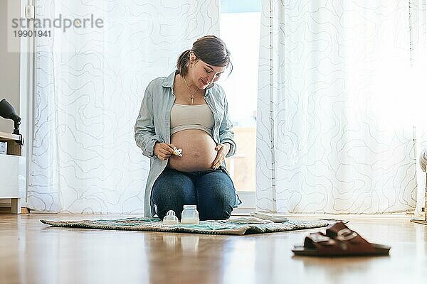 Glückliche kaukasische schwangere Mutter sitzt auf dem Boden  berührt ihren Bauch  Blue Jeans