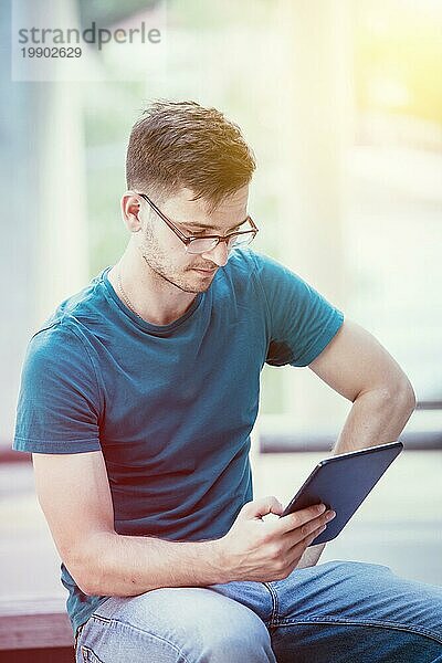 Hübscher junger Student sitzt im Freien auf dem städtischen Campus und arbeitet mit einem Tablet PC