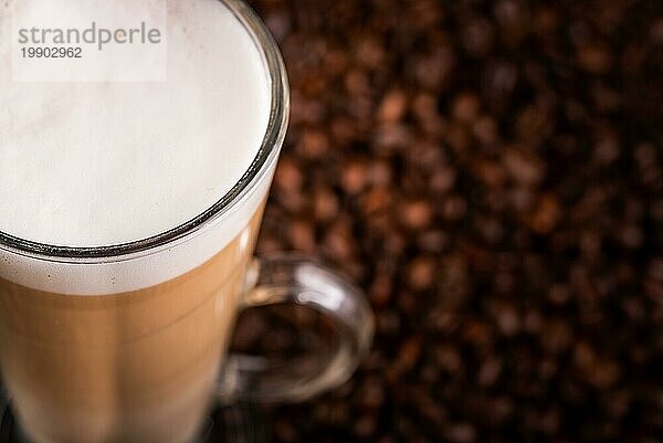 Milchkaffee mit schaumiger Milch in einem hohen Glas  auf Kaffeebohnen vor schwarzem Hintergrund. Ansicht von oben