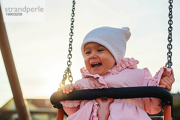 Adorable Baby Mädchen mit großen schönen Augen und eine Mütze mit Spaß auf einer Schaukel Fahrt auf einem Spielplatz in einem sonnigen Park