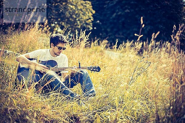 Junger Mann sitzt an einem Fluss und spielt auf seiner Westerngitarre