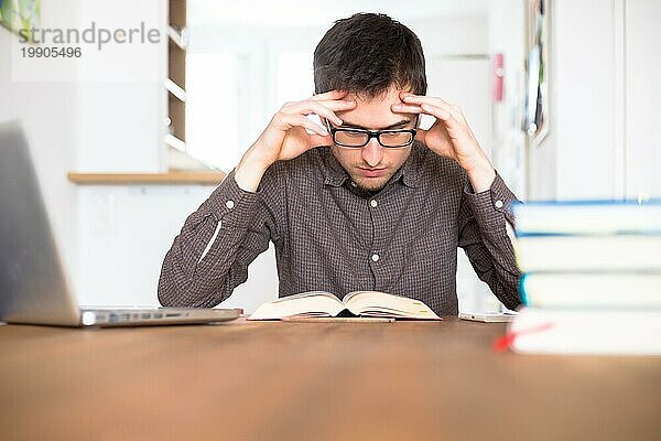 Männlicher erschöpfter und unzufriedener Student  der zu Hause recherchiert und lernt  Stapel von Büchern unscharf