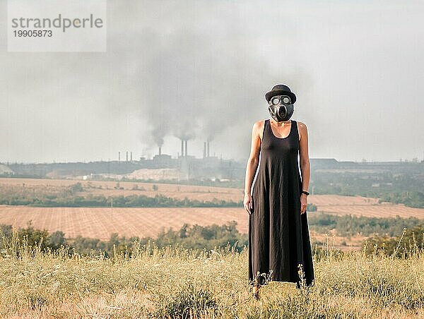 Junges Mädchen in einem schwarzen Kleid und einer Gasmaske vor dem Hintergrund rauchender Fabrikschornsteine