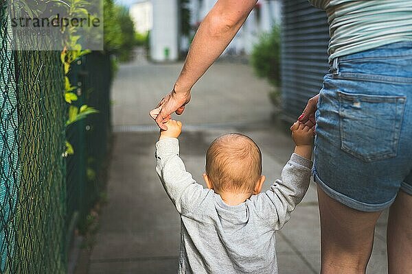 Konzept des Laufenlernens: Ein Kleinkind hält die Hand seiner Mutter und macht seine ersten Schritte. Nahaufnahme von hinten