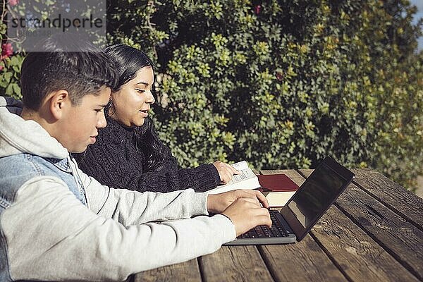 Zwei Kinder lernen mit einem Laptop und Büchern auf einem Holztisch im Freien an einem sonnigen Tag