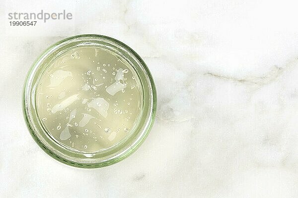 Ein Overhead Foto eines Glasgefäßes mit Aloe Vera Gel auf einem weißen Marmorhintergrund mit Platz für Text