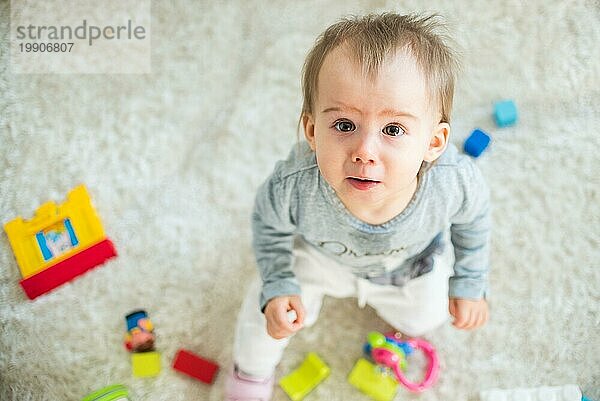 Porträt eines einjährigen Mädchens  das drinnen auf dem Teppich in einem hellen Raum spielt und nach oben schaut