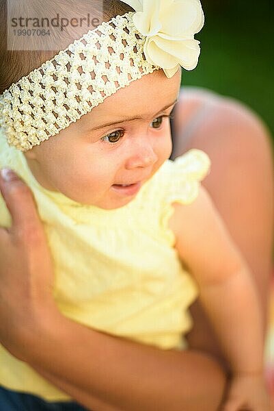 Portrait junge Mutter hält niedlichen Baby Mädchen in gelben Band und Kleid. Sonniger Sommertag im Freien. Mutterschaft Konzept