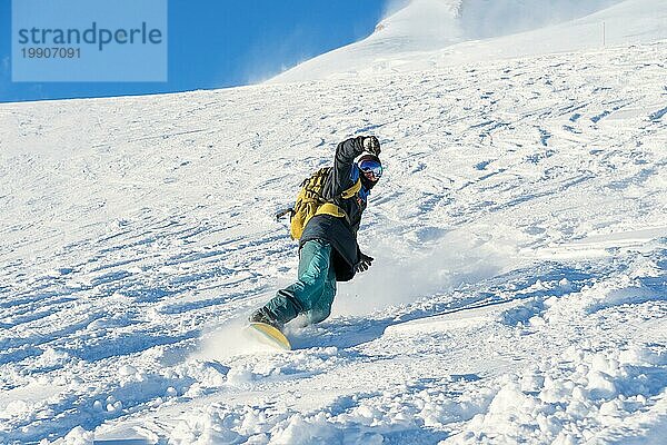 Ein Freeboard Snowboarder mit Skimaske und Rucksack fährt über die schneebedeckte Piste und hinterlässt einen Pulverschnee vor dem blaün Himmel