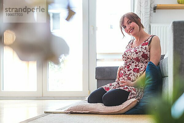 Schwangere kaukasische glückliche Mutter sitzt auf dem Boden  berührt ihren Bauch  blumiges Kleid