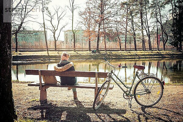 Rückenansicht einer jungen Frau mit Fahrrad  die die Sonne auf einer Parkbank genießt  Frühling