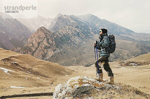 Ein Mann mit Bart und Sonnenbrille in einer Membranjacke  Hut  mit einem Rucksack und Stöcken für Nordic Walking  ein Reisender  der in der freien Natur steht und auf die Berge schaut