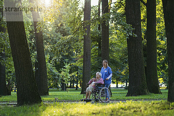 Lächelnde Mitarbeiterin im Gesundheitswesen geht mit einer älteren Frau spazieren  die im Rollstuhl in der Nähe von Bäumen im Park sitzt