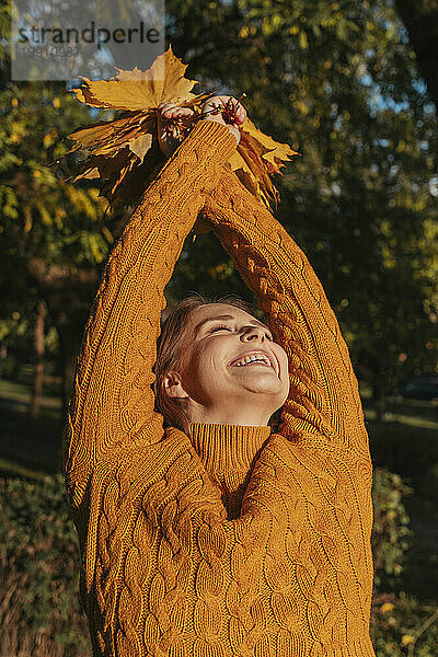 Fröhliche Frau mit erhobenen Armen und einem Bündel Ahornblätter im Herbstpark