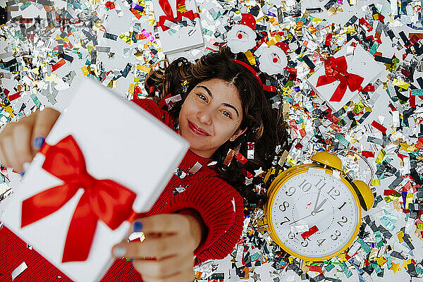 Lächelndes Mädchen hält Geschenkbox in der Hand und liegt auf Konfetti neben der Uhr
