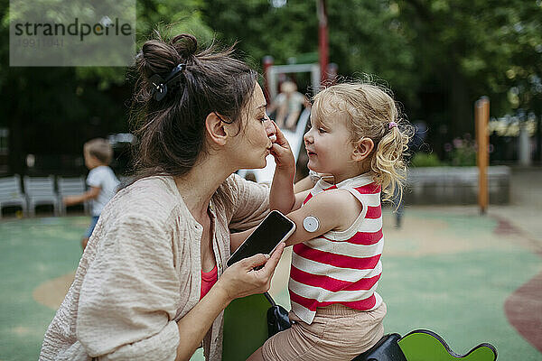 Mutter mit Smartphone schaut auf kleines Mädchen mit einem kontinuierlichen Glukosemonitor-Sensor auf ihrer Schulter