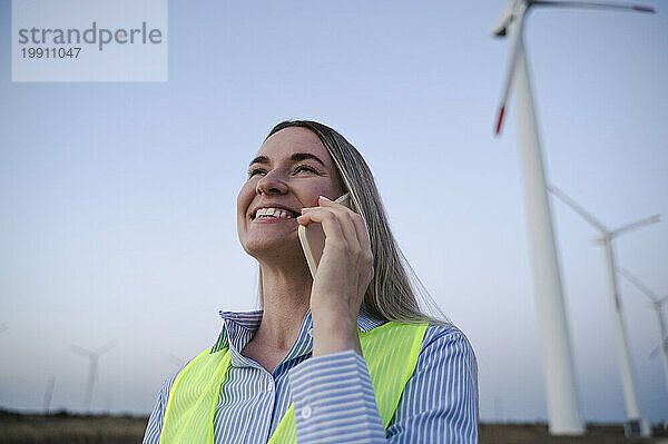Glücklicher Ingenieur  der in der Nähe von Windkraftanlagen mit dem Smartphone spricht