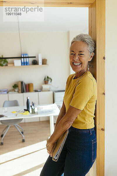 Happy mature businesswoman holding file folder and leaning on doorway in office