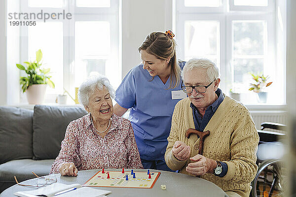 Fröhlicher Mitarbeiter im Gesundheitswesen mit älterem Paar  das am Tisch im Wohnzimmer Ludo-Spiel spielt