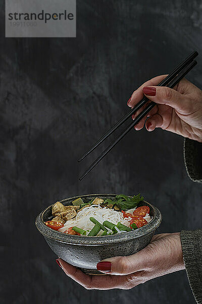 Hände einer Frau  die eine Schüssel mit veganer Tom-Kha-Kai-Suppe mit Tofu  Tomaten  Salat  Reisnudeln  Sesam und Frühlingszwiebeln isst