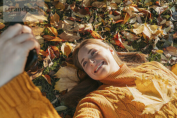 Lächelnde Frau liegt auf Blättern und macht ein Selfie im Herbstpark