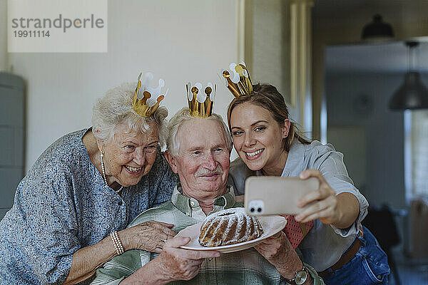 Fröhlicher Mitarbeiter im Gesundheitswesen  der ein Selfie mit einem älteren Paar macht  das Kuchen in der Hand hält und Geburtstag feiert