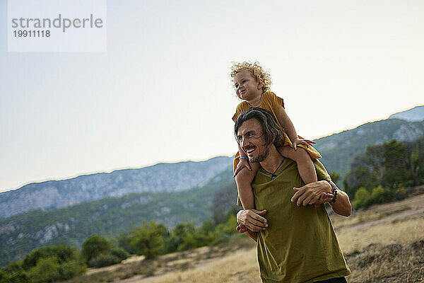 Glücklicher Mann  der seine Tochter auf Schultern trägt und vor den Bergen steht
