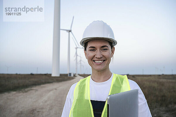 Glücklicher Ingenieur mit Schutzhelm  der mit Laptop vor Windkraftanlagen steht